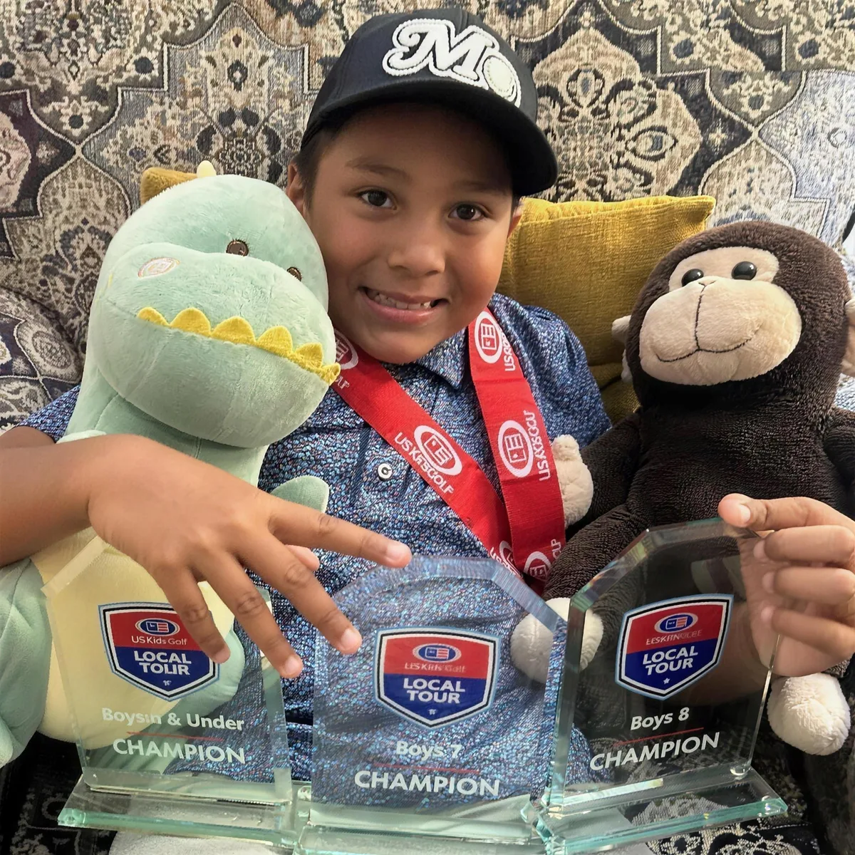 Junior golfer in a Mo hat holding three U.S. Kids Golf Local Tour champion trophies