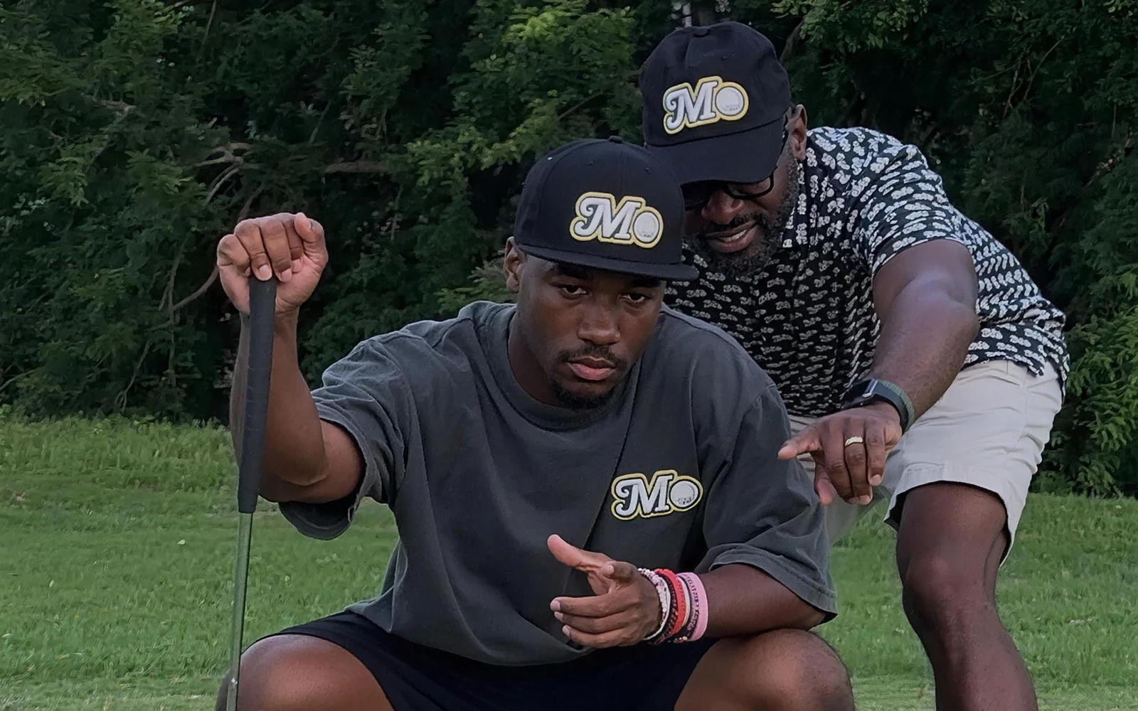 A coach in a Mo City Golf hat pointing out the line of a putt to a young golfer crouched on the green